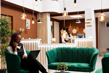 Young african american woman receptionist talking by phone while sitting with laptop at lounge...