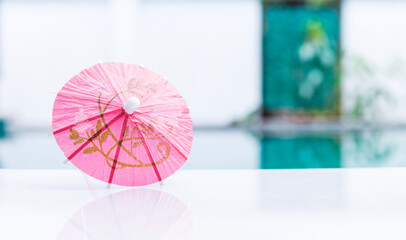 Pink paper umbrella on swimming pool edge with space on blurred background, summer outdoor day light, tropical summer concept