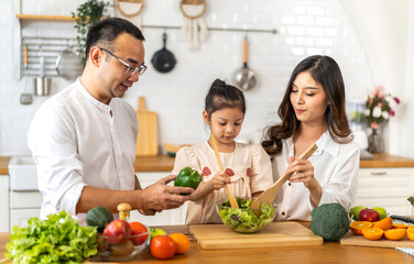 Naklejka premium Portrait of enjoy happy love asian family father and mother with little asian girl daughter child having fun help cooking food healthy eat together with fresh vegetable salad ingredient in kitchen