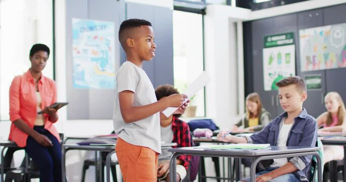 Diverse female teacher and happy schoolchildren at desks reciting in school classroom
