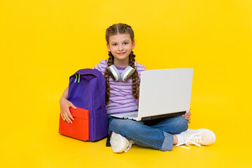 The child is engaged in an online lesson with a laptop. A young girl is sitting cross-legged on the floor with a backpack and headphones. Yellow isolated background.