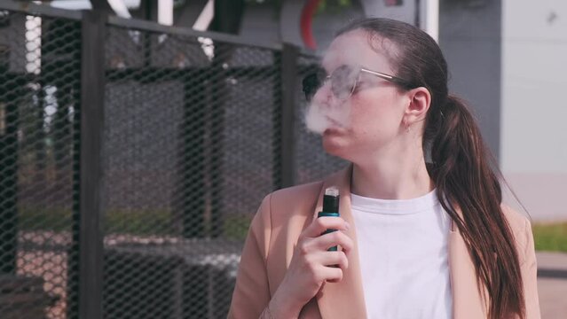 Beautiful girl in sunglasses and a classic jacket smokes a vape in a city park on a sunny day
