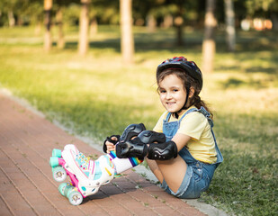 Cute little girl in protective equipment and rollers sitting on walkway in park outdoor. Summer...