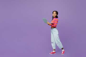 Full body side view little kid teen IT girl 15-16 years old wear striped orange sweatshirt hold use work on laptop pc computer walk go isolated on plain purple background. Childhood lifestyle concept.