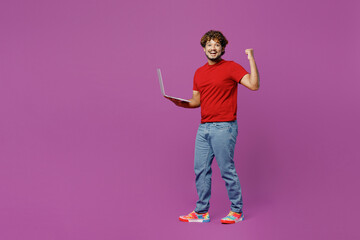 Full body side view young smiling happy Indian man wear red t-shirt casual clothes hold use work on laptop pc computer do winner gesture isolated on plain purple background studio. Lifestyle concept.