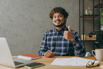 Successful smiling happy cheerful fun employee business Indian man he wearing casual blue checkered shirt looking camera show thumb up gesture sit work at office desk with laptop pc computer indoors.