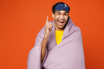 Calm young insighted man wear pyjamas jam sleep eye mask wrapped in purple blanket point finger up rest relax at home isolated on plain orange background studio portrait. Good mood night nap concept.