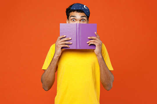 Calm young shocked amazed man he wear pyjamas jam sleep eye mask rest relax at home cover mouth with book look camera isolated on plain orange background studio portrait. Good mood night nap concept.