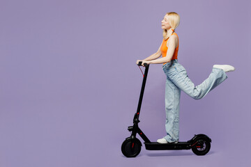 Full body sideways young blonde woman wear orange tank shirt casual clothes riding electric scooter raise up leg isolated on plain pastel light purple background studio portrait. Lifestyle concept. © ViDi Studio