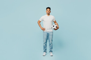 Full body smiling young man fan wear t-shirt cheer up support football sport team hold in hand soccer ball stand akimbo look camera watch tv live stream isolated on plain pastel blue color background.