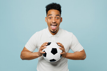 Young excited overjoyed man fan wearing basic t-shirt looking camera cheer up support football sport team hold in hand soccer ball watch tv live stream isolated on plain pastel blue color background.