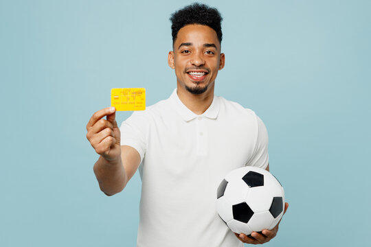 Young happy man fan wears basic t-shirt cheer up support football sport team hold in hand mock up of credit bank card soccer ball watch tv live stream isolated on plain pastel blue color background.