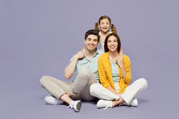 Full body smiling happy fun young happy parents mom dad with child kid daughter girl 6 years old wear blue yellow casual clothes sit hug cuddle isolated on plain purple background. Family day concept.