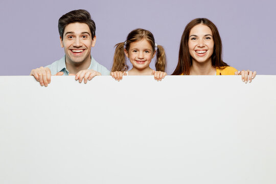 Young smiling parents mom dad with child kid daughter girl 6 years old wearing blue yellow casual clothes hold big white empty poster billboard isolated on plain purple background. Family day concept.
