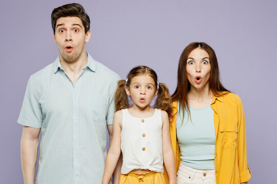 Young Shocked Happy Parents Mom Dad With Child Kid Daughter Girl 6 Years Old Wear Blue Yellow Casual Clothes Looking Camera With Opened Mouth Isolated On Plain Purple Background. Family Day Concept.