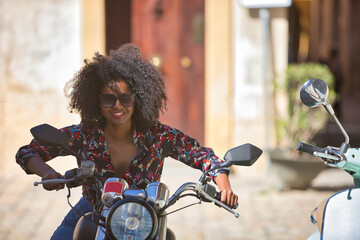 Fototapeta na wymiar Young woman, beautiful and black with afro hair, with floral shirt and sunglasses, mounted on a motorcycle in a happy and fun attitude. Motorcycle concept, fun, transport.