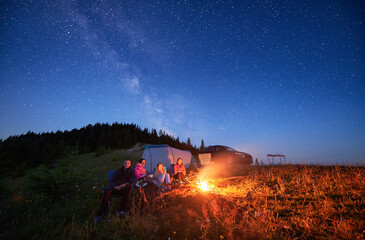 Family camp in mountains in the evening. Group of tourists having a rest near campfire on a top of hill, near tourist tent and off-road vehicle. Starry night in Carpathian mountains.