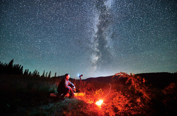 Man taking photos of starry sky with Milky way. Male photographer sitting on log and focusing camera. Man with camera sitting next to camp fire in mountains.