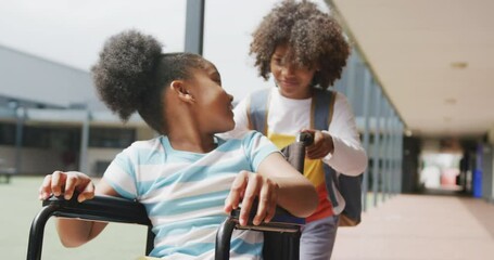 Video of two happy diverse schoolchildren, boy pushing girl in wheelchair in corridor - Powered by Adobe