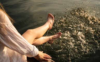 A woman relaxes by the lake, sitting on the edge of a wooden jetty, swinging her legs of the water.
