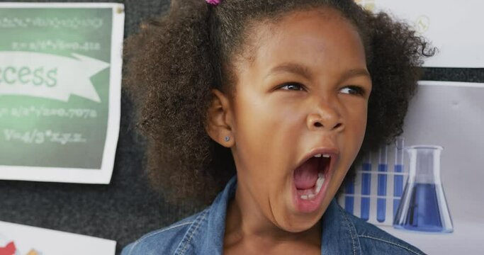 Video portrait of happy, tired biracial schoolgirl yawning in school classroom