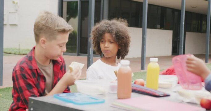 Video of two diverse schoolboys eating packed lunch and talking in schoolyard