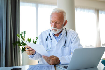 Serious old mature professional male doctor using laptop computer in hospital office having medical...