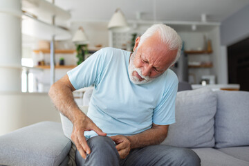 Older man sitting on a home couch, holding his leg in pain due to knee joint discomfort, the...