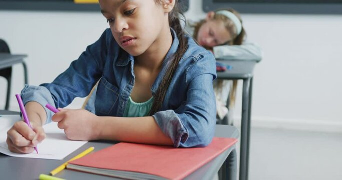 Video of bored biracial schoolgirl sitting at desk writing in class, copy space