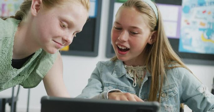 Video of two happy caucasian schoolgirls looking at tablet and talking in classroom, copy space