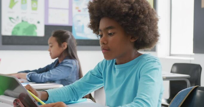 Video of concentrating african american schoolboy sitting at desk using tablet in class, copy space