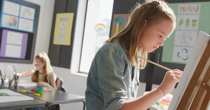 Video of concentrating caucasian schoolgirl standing at easel painting in art class, copy space