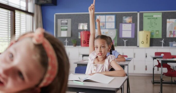 Happy diverse schoolgirls raising hands at desks in elementary school class, slow motion