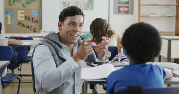 Happy diverse male teacher talking to schoolboy at desk in elementary school class, slow motion