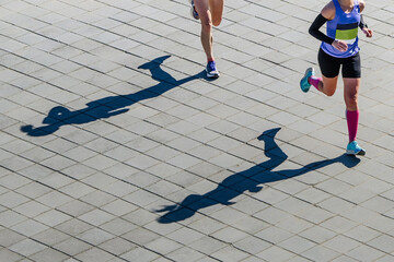 overhead view two female athletes running marathon race, shadows of runners on paving slabs