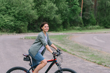 Satisfied smiling happy woman in casual green jacket jeans riding bicycle bike on sidewalk in park outdoors, look camera. People active urban healthy lifestyle cycling concept.