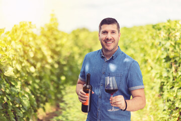 Smiling young man holding glass and bottle of wine in vineyard