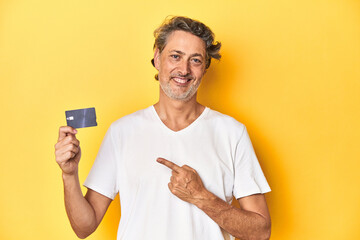 Man holding a credit card, yellow studio backdrop smiling and pointing aside, showing something at blank space.