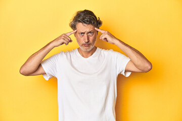 Middle-aged man posing on a yellow backdrop focused on a task, keeping forefingers pointing head.