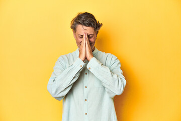 Middle-aged man posing on a yellow backdrop holding hands in pray near mouth, feels confident.