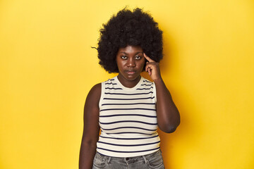 Fototapeta na wymiar African-American woman with afro, studio yellow background pointing temple with finger, thinking, focused on a task.