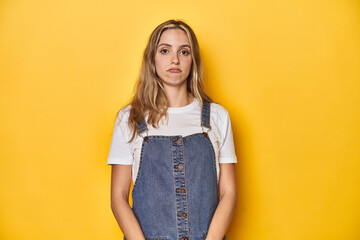 Young blonde Caucasian woman in denim overalls posing on a yellow background, sad, serious face, feeling miserable and displeased.