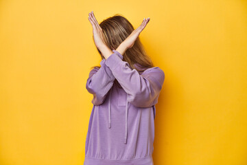 Young blonde Caucasian woman in a violet sweatshirt on a yellow background, keeping two arms...