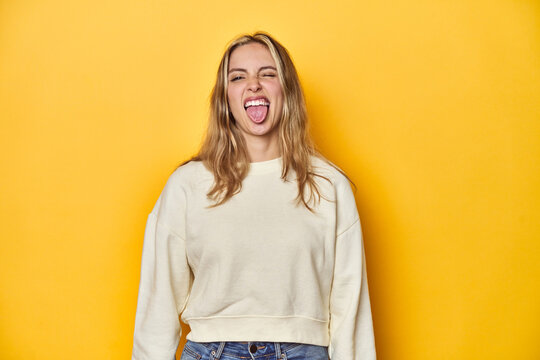 Young Blonde Caucasian Woman In A White Sweatshirt On A Yellow Studio Background, Funny And Friendly Sticking Out Tongue.