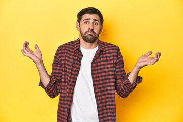 Caucasian man in red checkered shirt, yellow backdrop doubting and shrugging shoulders in...