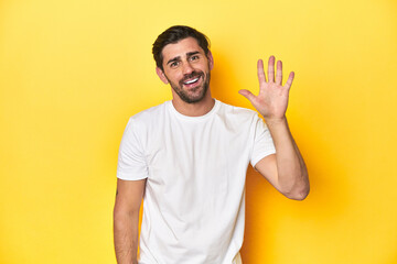Caucasian man in white t-shirt on yellow studio background smiling cheerful showing number five...