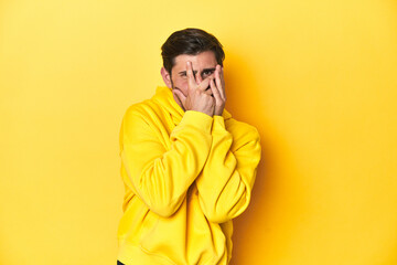 Man in yellow hoodie, monochrome studio backdrop blink through fingers frightened and nervous.