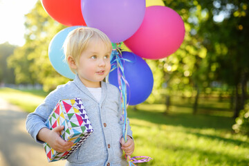 Cute little child going to congratulate a friend on his birthday. Toddler holding bundle of colorful balloons and gift in a festive box. Anticipation of the holiday.