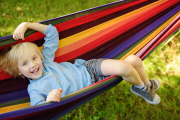 Fototapeta na wymiar Cute little blond caucasian boy relaxing and having fun in multicolored hammock in backyard or outdoor playground. Summer active leisure for kids. Child swinging on hammock.