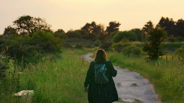 The serene countryside sets the stage as a young woman, backpack in tow, leisurely traverses a picturesque path. Surrounding trees and vibrant plants accompany her slow-motion journey.